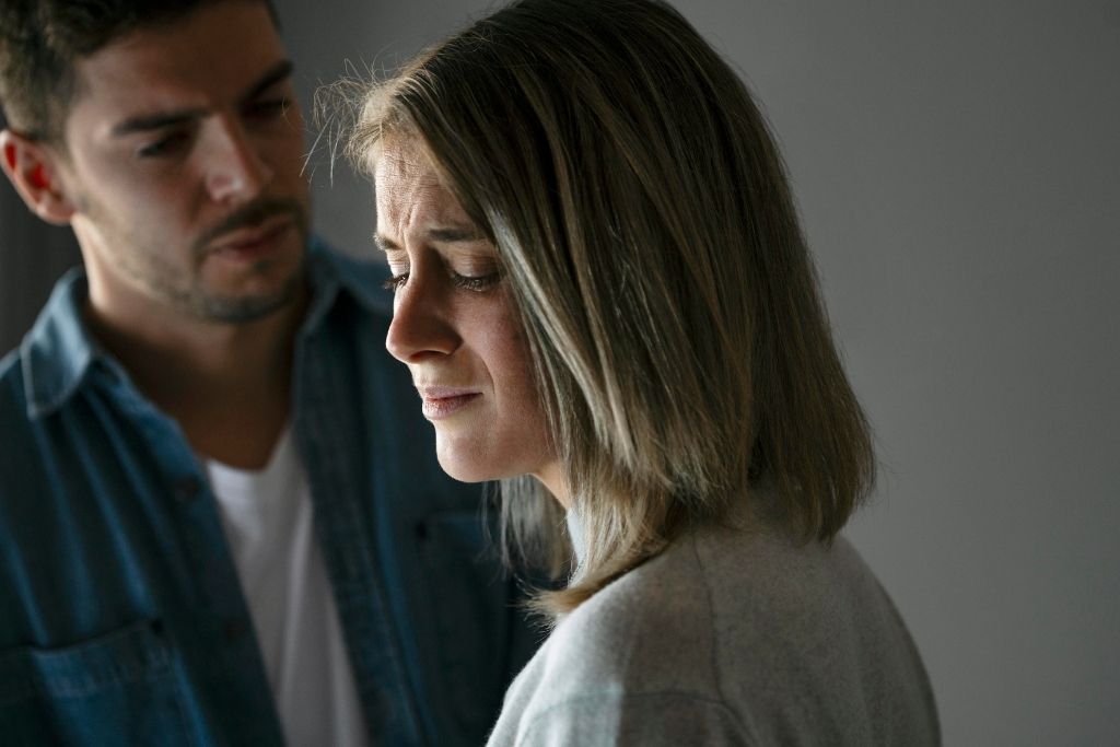 Apego ansioso en pareja: mujer triste y preocupada mientras su pareja la observa de fondo.