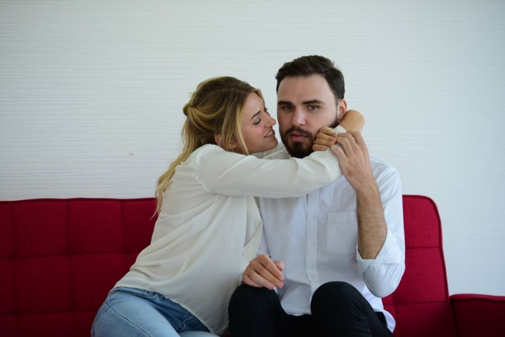 Apego ansioso en pareja: mujer abraza a un hombre en un sofá rojo mientras él se muestra incómodo.